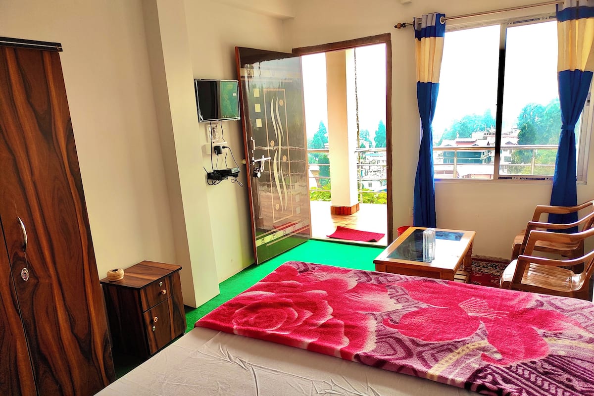 A room with a bed, a wooden wardrobe, a small table, a chair, a TV mounted on the wall, and a view of the outside through a glass door.