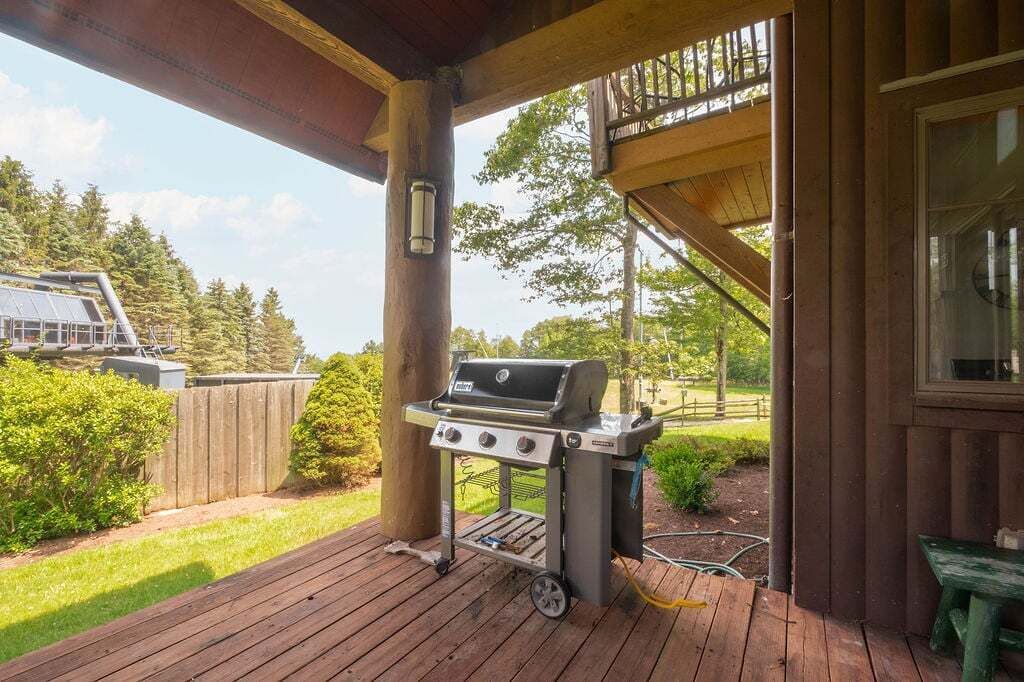 A covered outdoor area with a barbecue grill, wooden deck, and a view of a forested area.