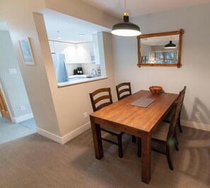 Comedor con mesa de madera, sillas, una lámpara colgante y una cocina visible a través de una puerta abierta.