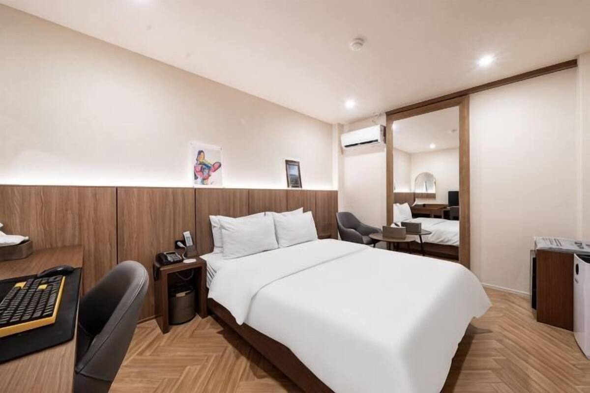 A hotel room with a large bed, a desk with a computer, a chair, a small table, and a trash bin.