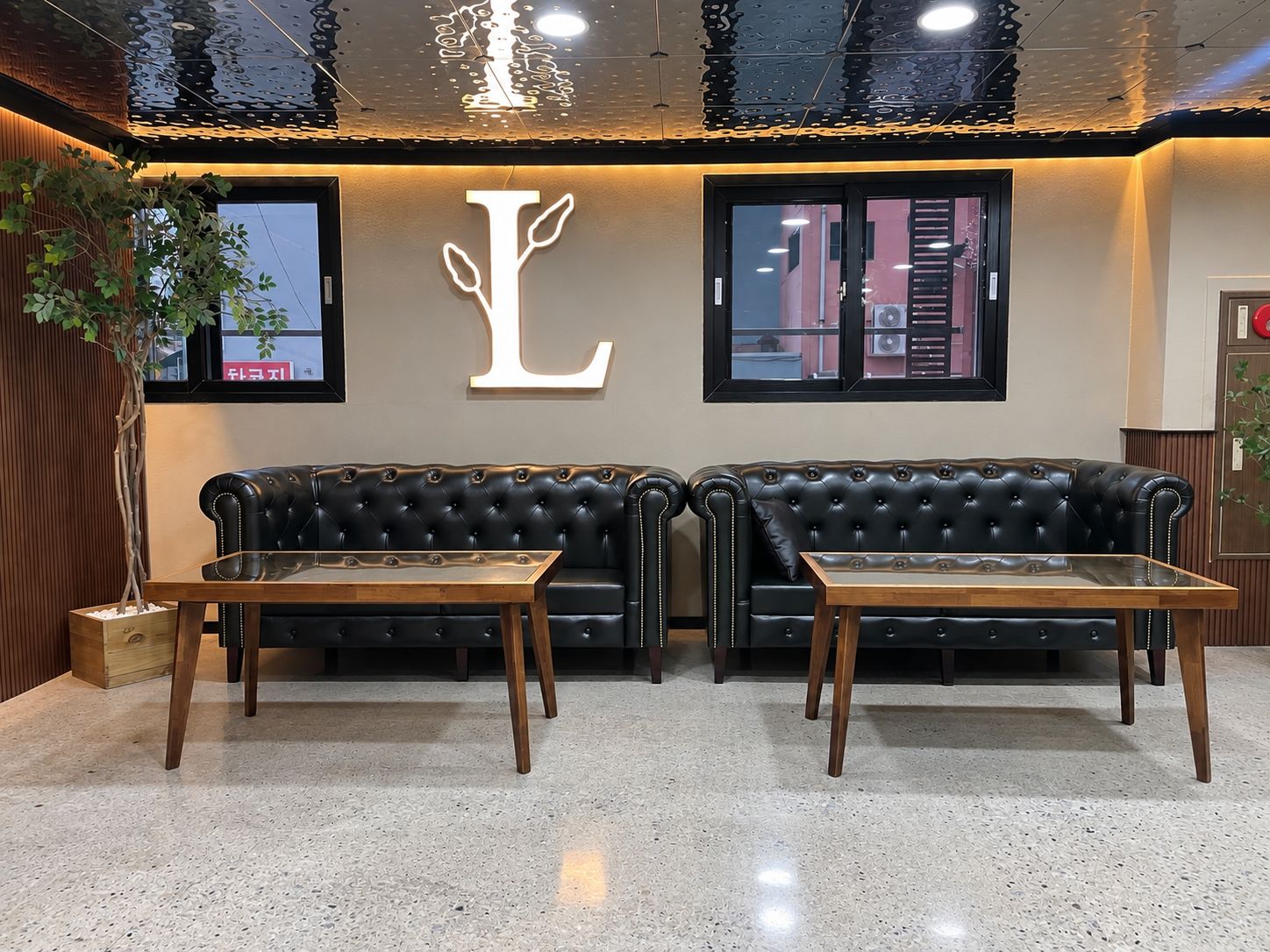 A lobby with two black leather sofas, wooden tables, a large illuminated letter 'L', and a plant.