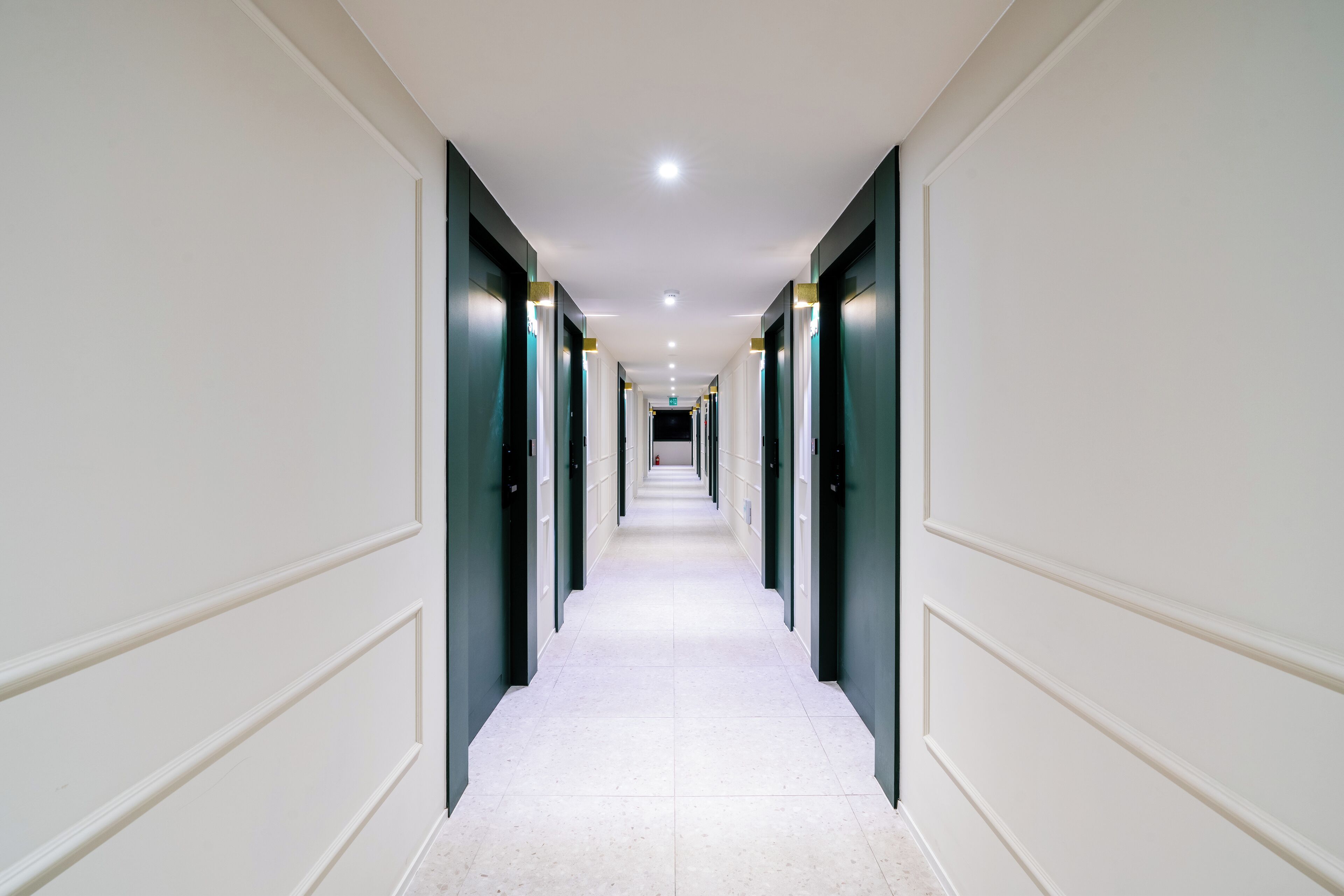 Un long couloir étroit aux murs blancs et aux portes vert foncé, qui mène à une sortie lumineuse au bout.