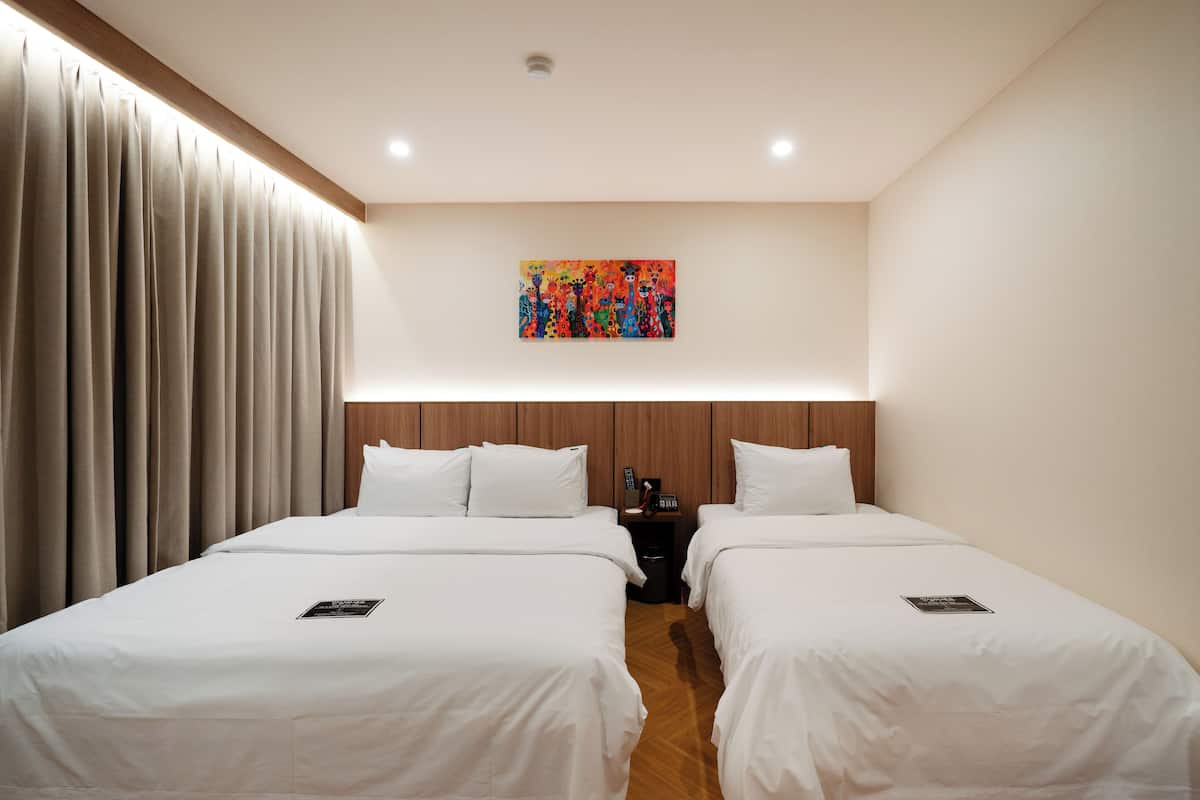 A hotel room with two beds, a wooden headboard, a painting on the wall, and a nightstand with a phone and a lamp.