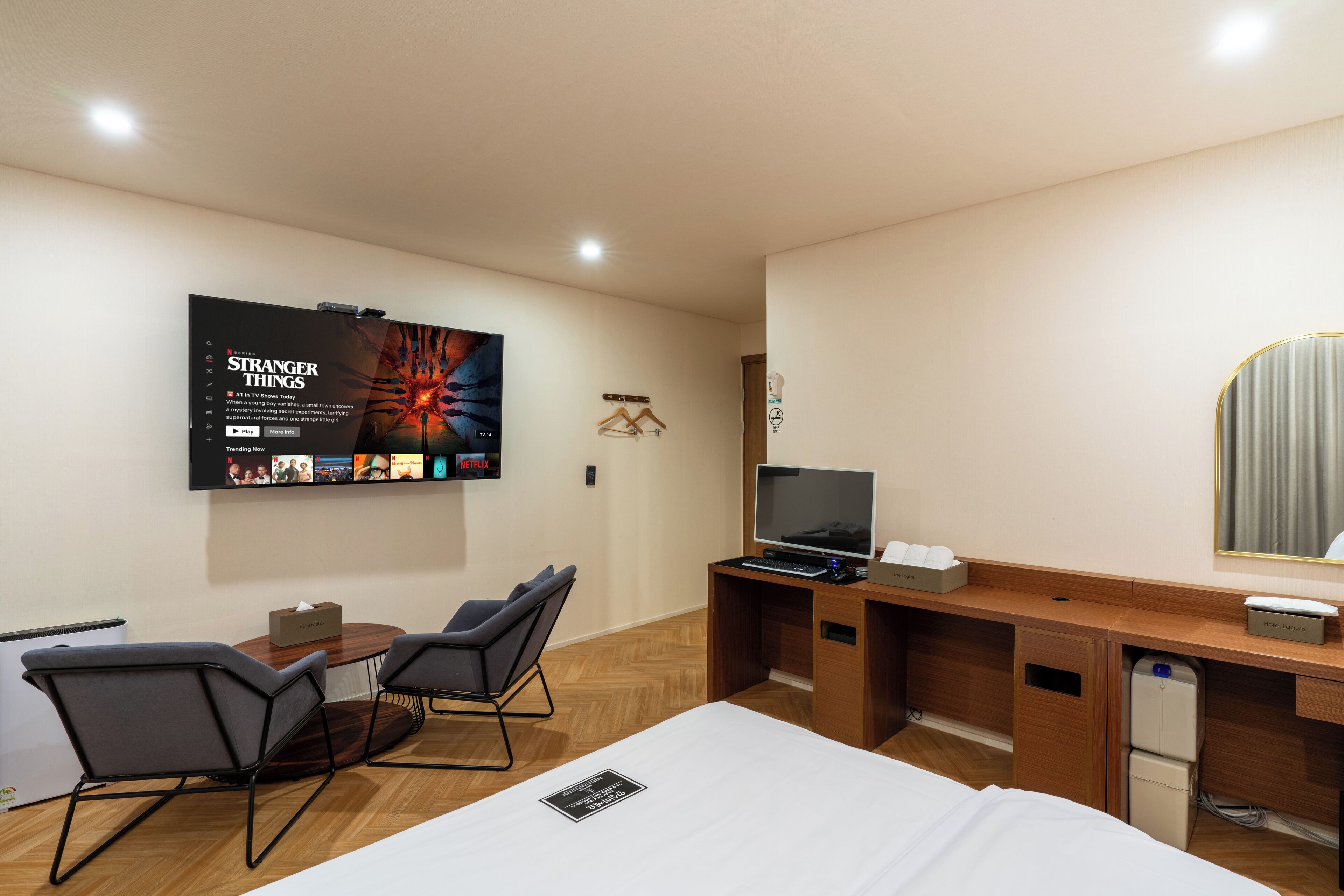 A hotel room with a bed, a desk with a computer, a TV, and a wall-mounted screen displaying 'Stranger Things'.