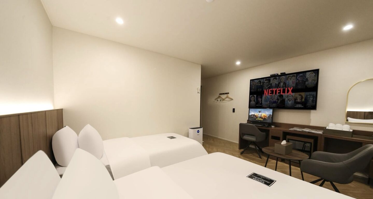 Habitación de hotel con dos camas, un escritorio, una silla, un televisor montado en la pared y una mesita.