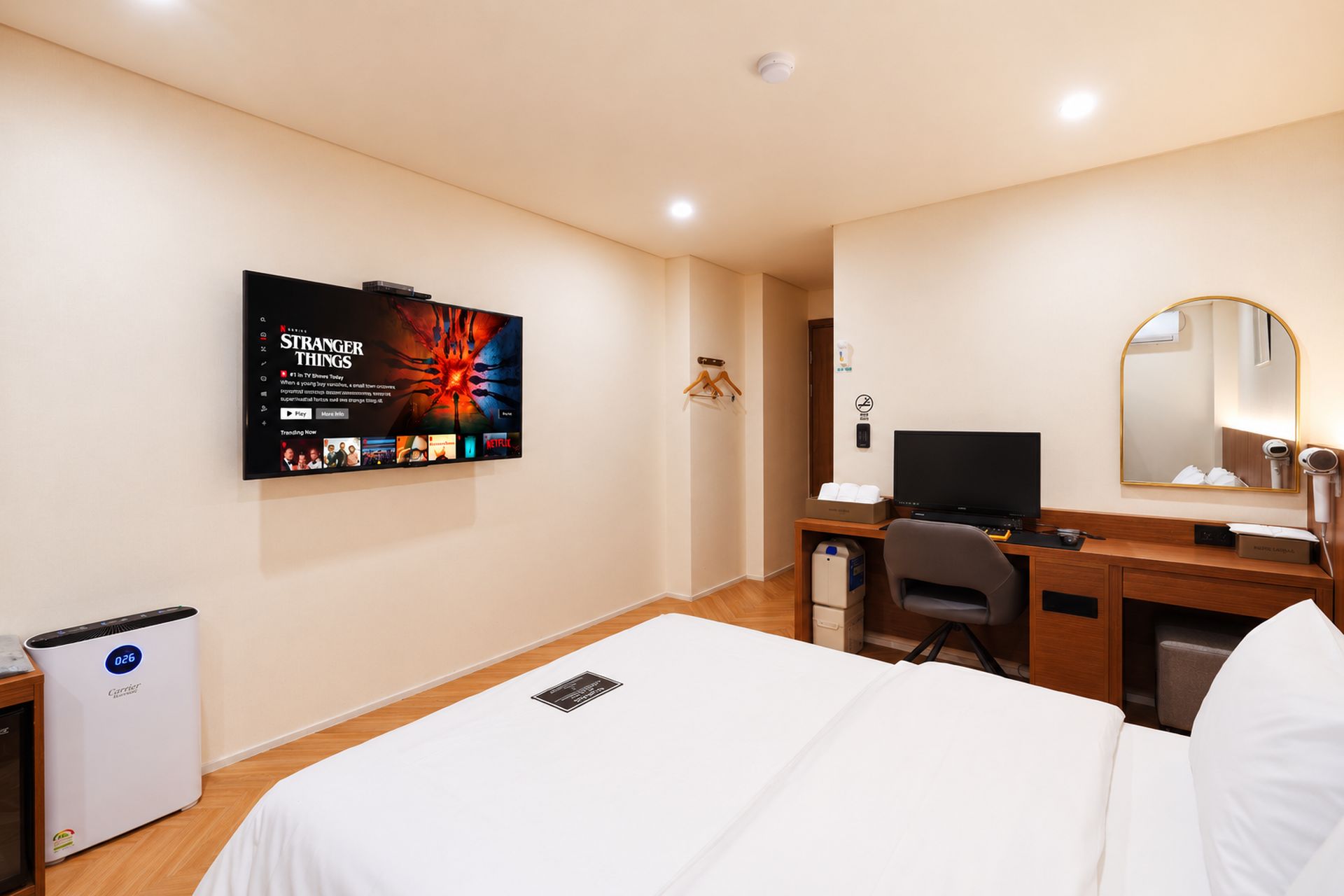 A hotel room with a bed, a desk with a computer, a TV mounted on the wall, a mirror, and a heater.