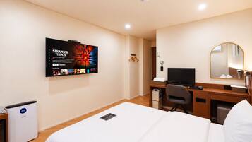 A hotel room with a bed, a desk with a computer, a TV mounted on the wall, a mirror, and a heater.