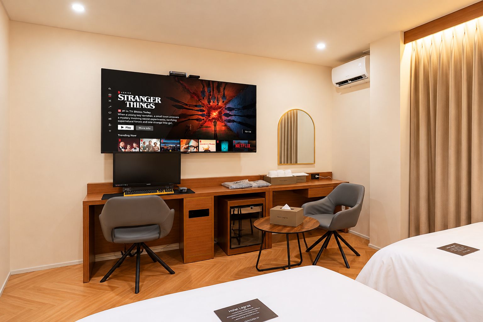 A hotel room with a TV displaying 'Stranger Things', a desk with a computer, two chairs, a small table with a tissue box, and a bed with a pillow.