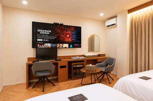 Habitación de hotel con una televisión mostrando 'Stranger Things', un escritorio con una computadora, dos sillas, una mesita con una caja de pañuelos y una cama con un almohadón.
