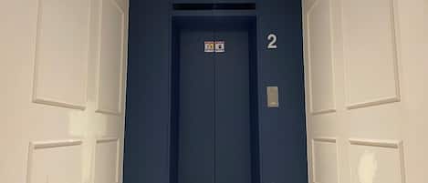 En hall med en blå hiss, vita väggar och ett klinkergolv.