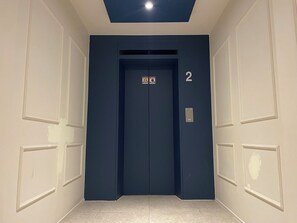 A hallway with a blue elevator, white walls, and a tiled floor. - Hotel Laglas Asan (Asan)
