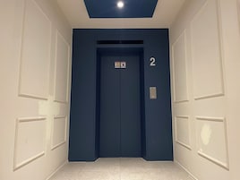 Un pasillo con un ascensor azul, paredes blancas y suelo de baldosas.