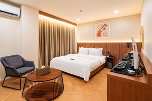 Una habitación de hotel con una cama, un escritorio con computadora, una silla, una mesita de noche con lámpara y una cortina.