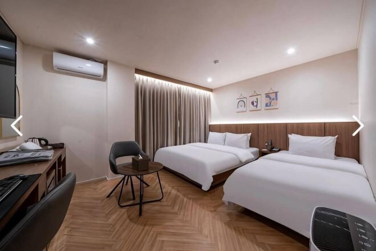 A hotel room with two beds, a desk with a chair, a TV, and an air conditioning unit.