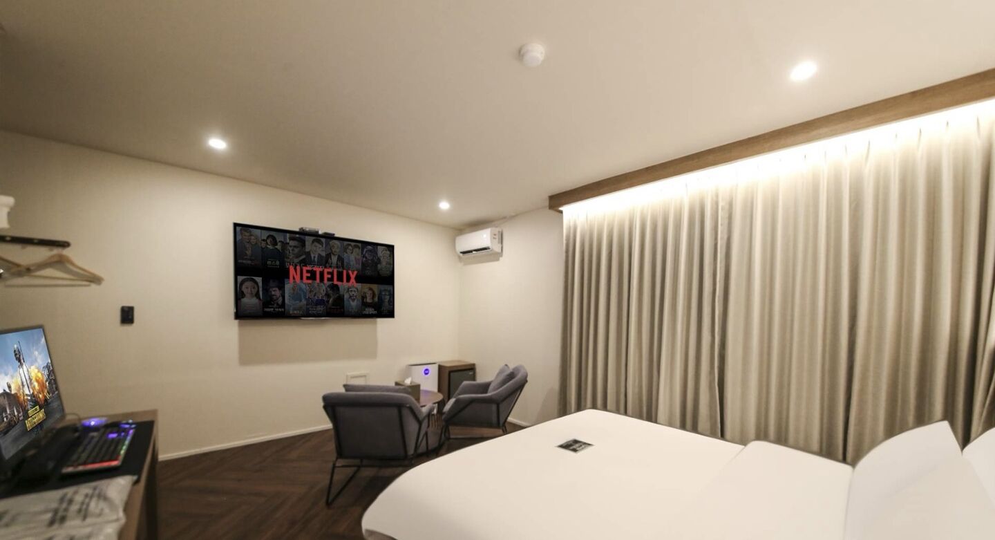 Ein Hotelzimmer mit einem Bett, einem Schreibtisch mit Computer, einem Sessel, einem an der Wand montierten Fernseher und einem Fenster mit Vorhängen.