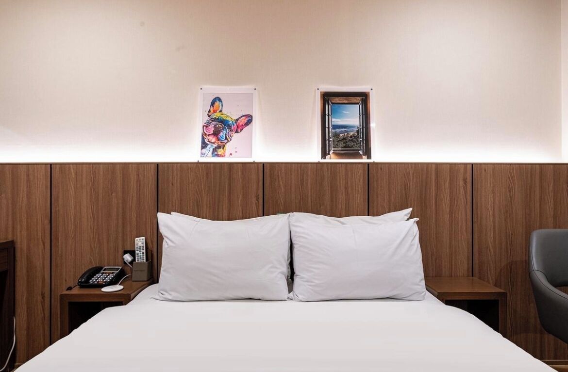 A hotel room with a wooden headboard, a bed with white linens, a bedside phone, a small table with a lamp, and two framed pictures on the wall.