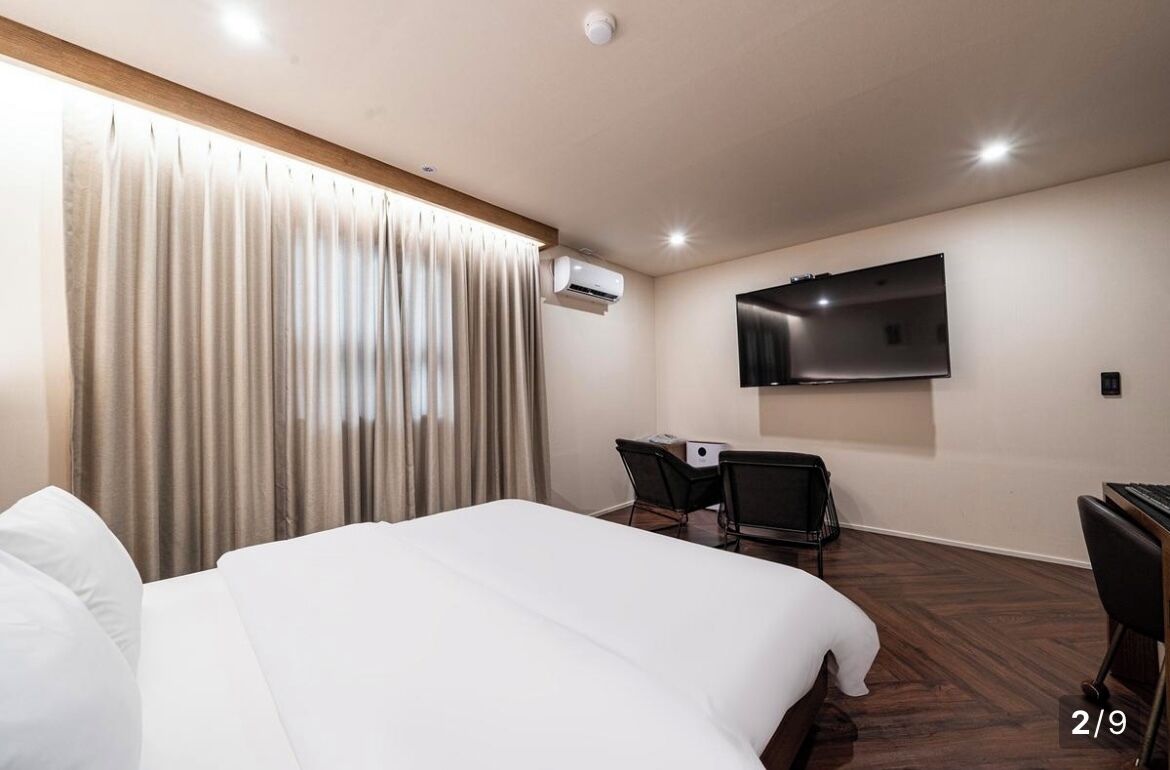 A hotel room with a large bed, a desk with a chair, a flat-screen TV mounted on the wall, and an air conditioning unit.
