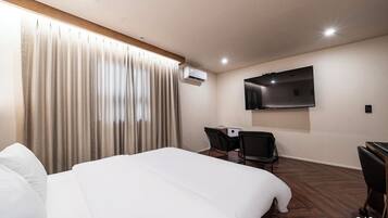 A hotel room with a large bed, a desk with a chair, a flat-screen TV mounted on the wall, and an air conditioning unit.
