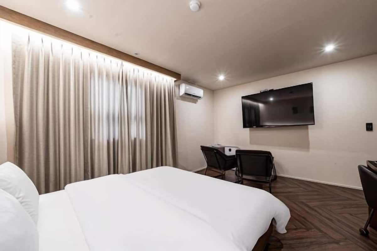 A hotel room with a bed, a desk, a chair, a TV, and an air conditioning unit.