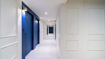Un lungo e stretto corridoio con porte alternate blu e bianche, illuminazione a incasso e pavimento piastrellato.