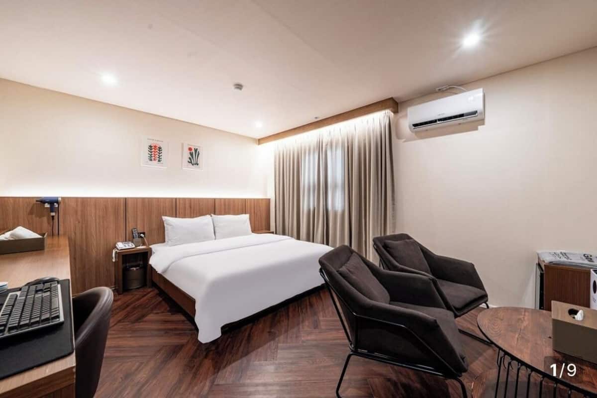 A hotel room with a bed, a desk with a computer, a chair, a small table, and an air conditioning unit.