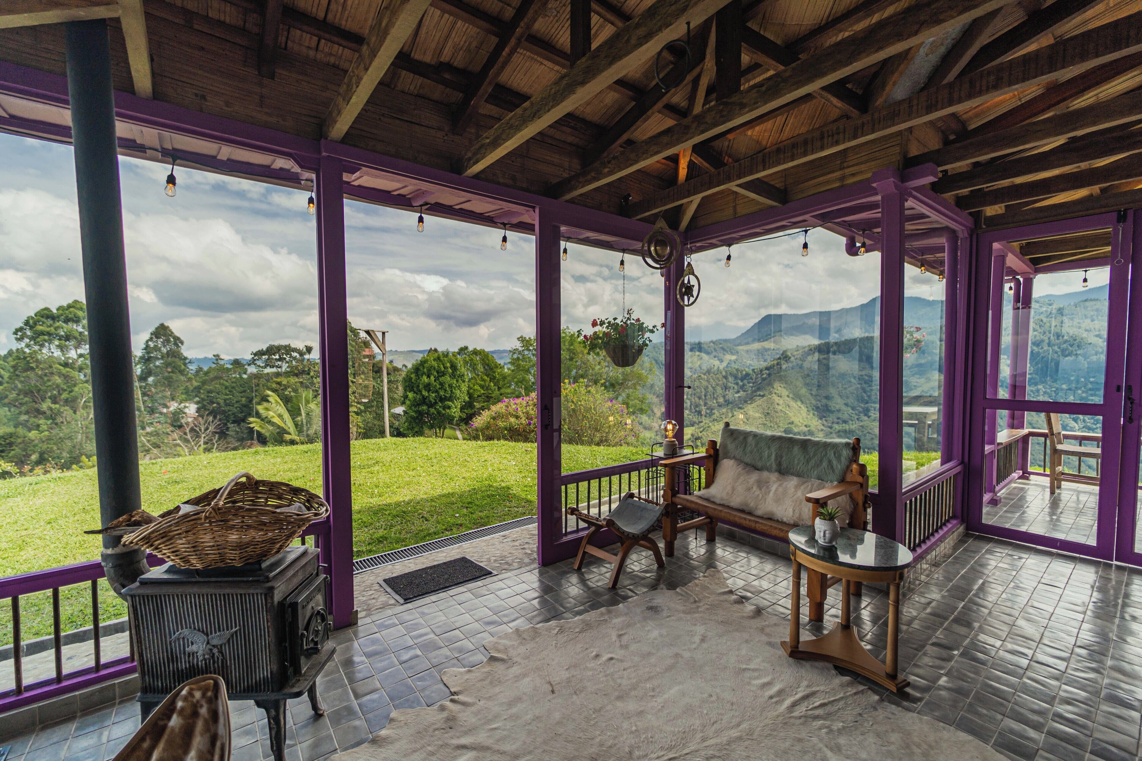 Un espacio al aire libre cubierto con techo de madera, barandilla morada, un banco, una mesa y vista a un paisaje verde.