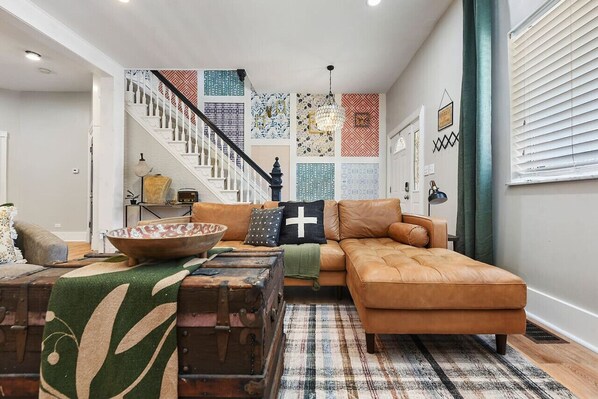 A living room with a brown leather sofa, a patterned rug, a vintage suitcase, and a staircase with patterned walls.