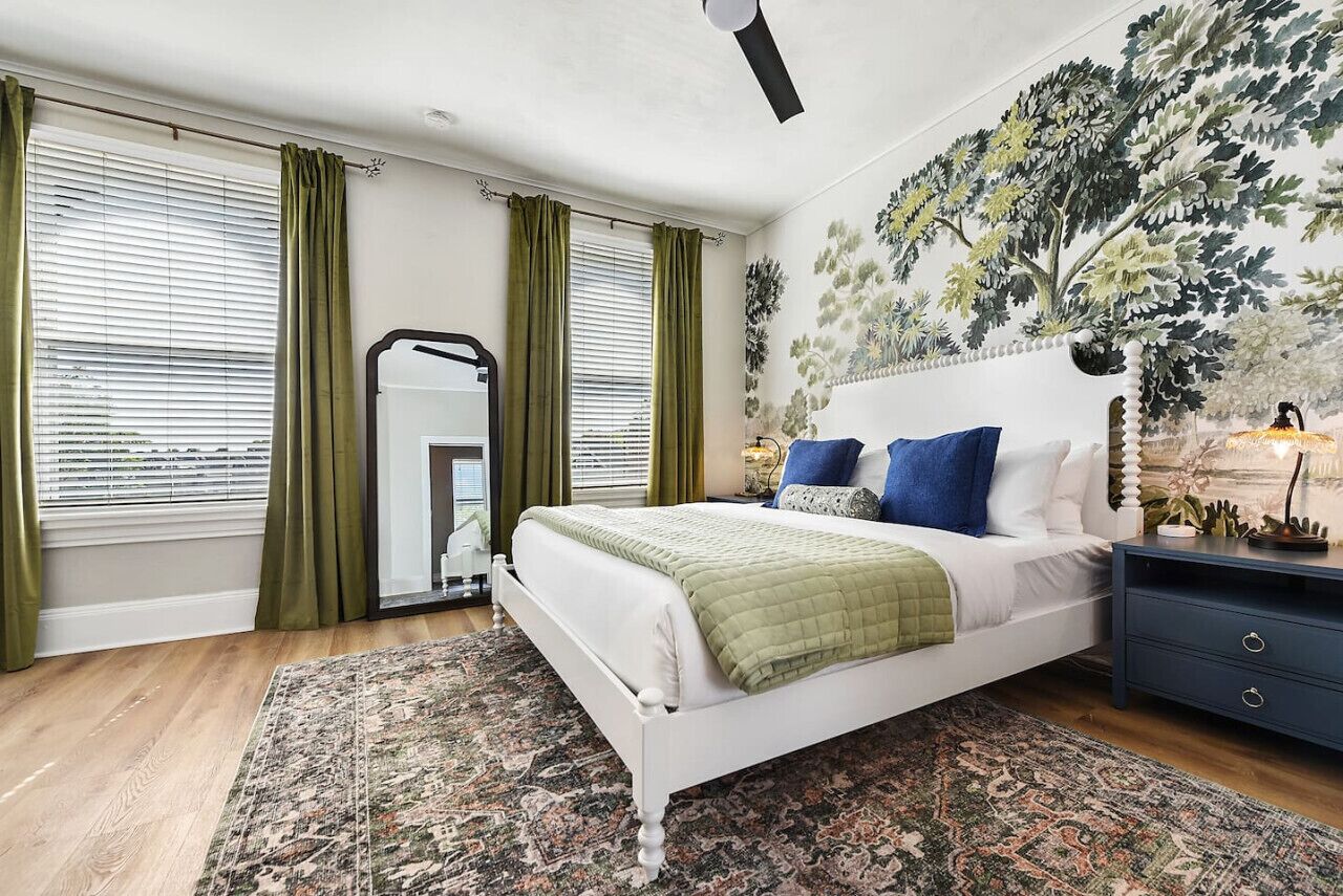 Ein Schlafzimmer mit Bett, grünen Vorhängen, einem großen Spiegel, einem Nachttisch und einem Wandbild.
