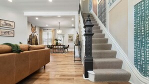 A spacious living room with a brown leather sofa, a dining area with a chandelier, and a staircase with patterned wallpaper. - ★ HostWise Stays ★ Foxy home, off-street parking, new remodel! (Pittsburgh)