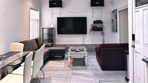A modern living room with a glass dining table, a sofa, a red couch, a ceiling fan, and a mounted TV.