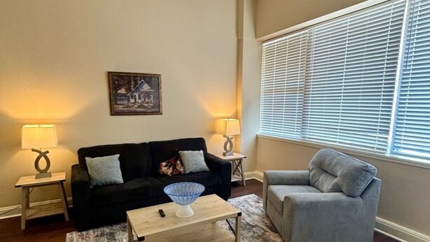 A living room with a black sofa, a grey armchair, a coffee table, and two lamps. There is a painting on the wall and a window with blinds. - Legacy BnB Suites (New Orleans)