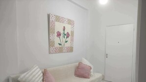 Een witte bank met twee roze en witte kussens, een bloemistisch kunstwerk aan de muur, en een witte deur.