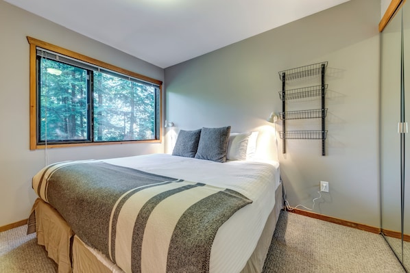 A bedroom with a large bed, a window with a view of trees, a bedside table with a lamp, and a wall-mounted shelf.