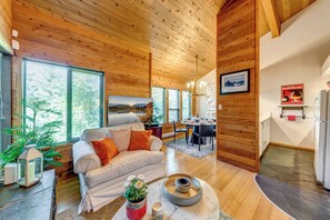 A cozy living room with a wooden ceiling, a sofa with orange cushions, a dining area with a table and chairs, and a kitchen area with a refrigerator.