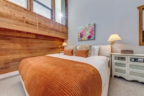Une chambre à coucher avec une tête de lit en bois, un grand lit recouvert d’une couverture marron, deux lampes de chevet, une table de chevet avec des tiroirs et un tableau floral accroché au mur.
