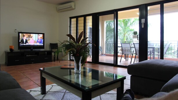 A living room with a flat-screen TV, a glass coffee table, and a potted plant. - Zen Bridgeport Waterfront Balcony Dining - Pool (Darwin)