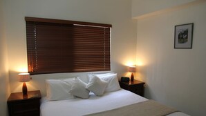 A neatly made bed with white linens and pillows, flanked by two bedside tables with lamps, a window with blinds, and a framed picture on the wall. - Zen Bridgeport Waterfront Balcony Dining - Pool (Darwin)
