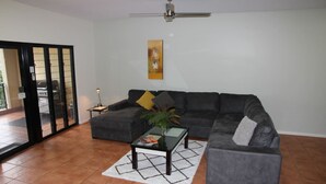 A living room with a gray sectional sofa, a glass coffee table, and a white rug. There is a ceiling fan, a painting on the wall, and a door leading to an outdoor area. - Zen Bridgeport Waterfront Balcony Dining - Pool (Darwin)