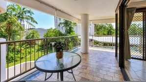 A balcony with a glass table and a potted plant, surrounded by a metal railing, overlooking a lush green area with palm trees. - Downtown Delight 2BR Getaway Pool (Darwin)