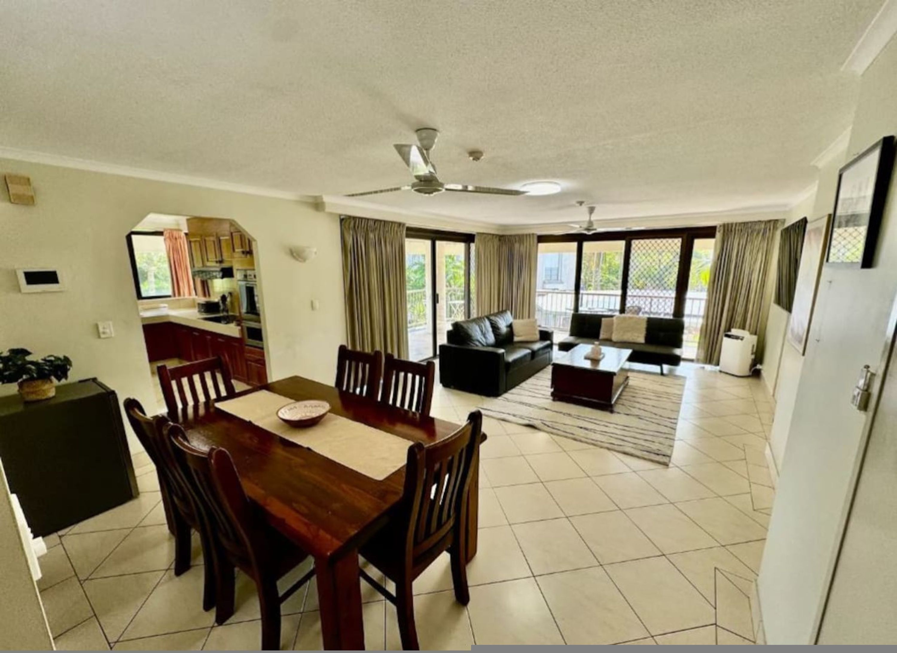 A spacious living area with a dining table and chairs, a sofa, and a coffee table. There is a ceiling fan and large windows with curtains.