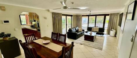 A spacious living area with a dining table and chairs, a sofa, and a coffee table. There is a ceiling fan and large windows with curtains.