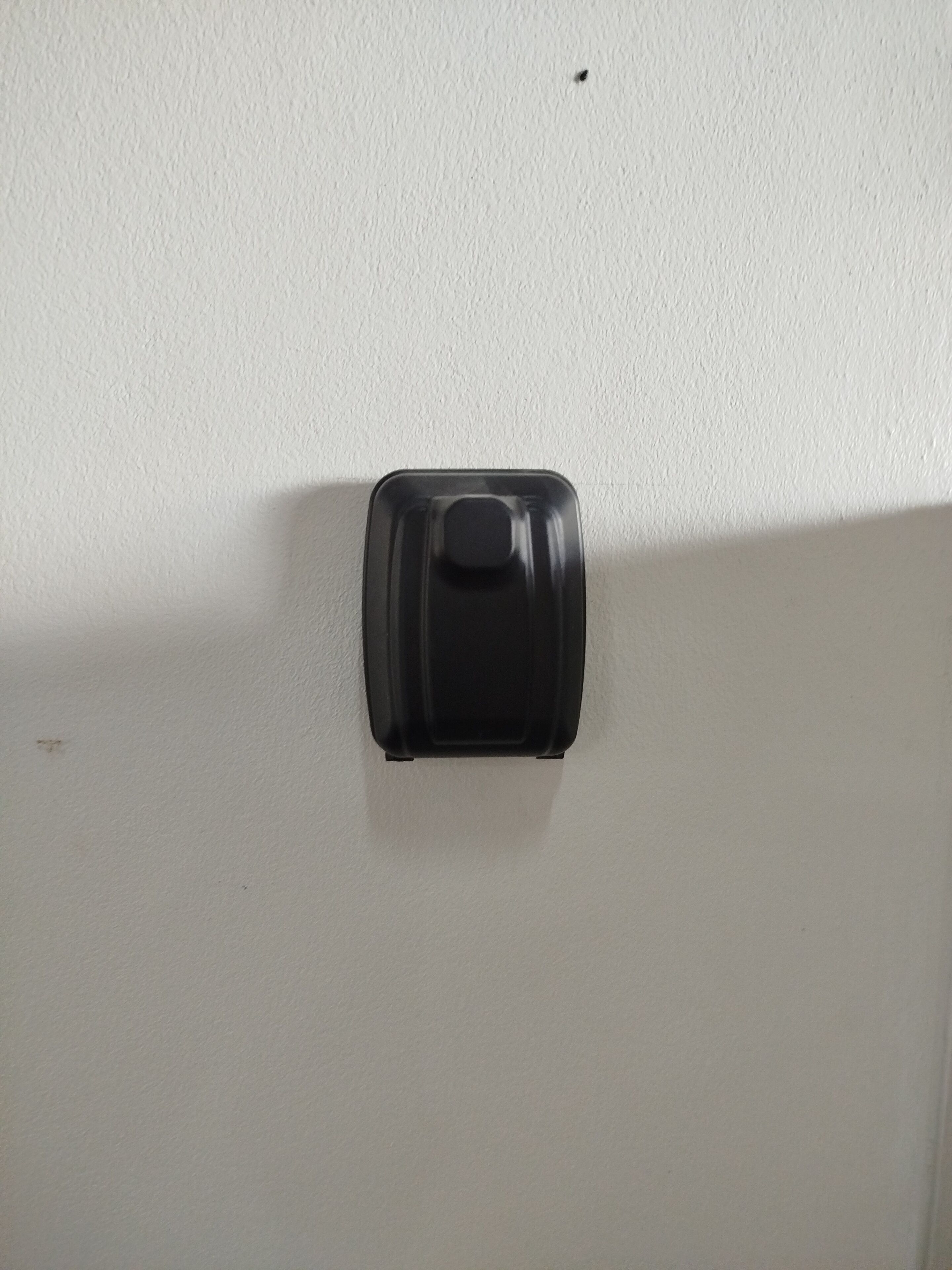 A white wall with a black object mounted on it, possibly a light switch or a control panel.