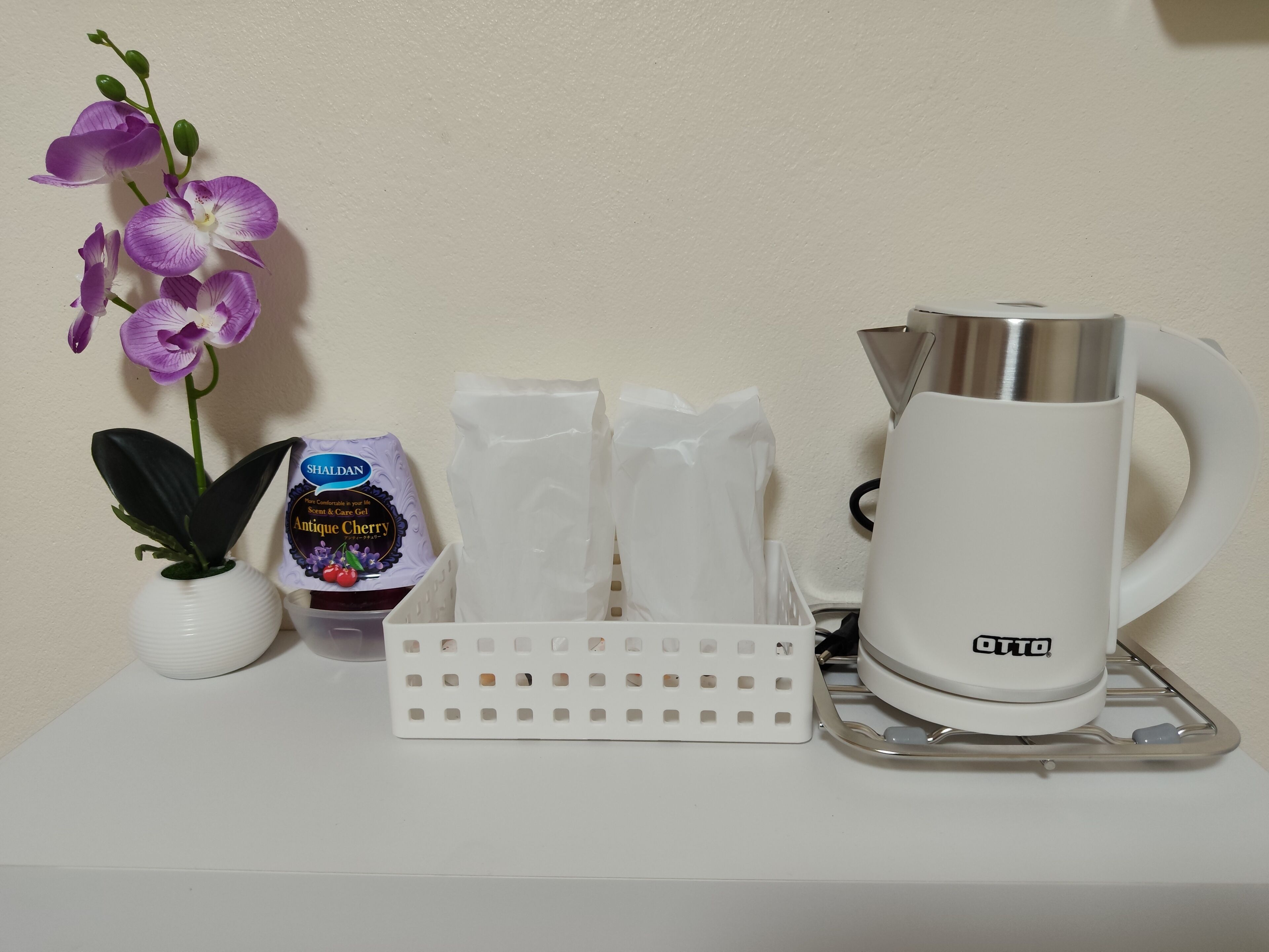 A white table with a potted orchid, a bottle of Shaldan Antique Cherry Care Gel, a white plastic tissue holder, and a white OTTO coffee maker.