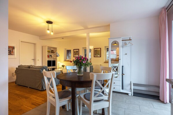 A dining area with a round table, white chairs, a vase with flowers, and a grey sofa in the background.