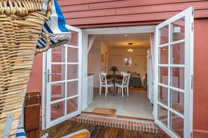 An open door leading to a dining area with a table and chairs, a wicker chair outside, and a red exterior wall.