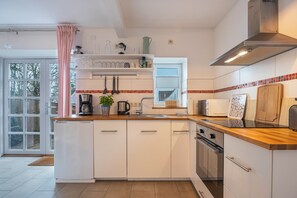 A modern kitchen with white cabinetry, a wooden countertop, a stainless steel range hood, and a window with a view of trees.