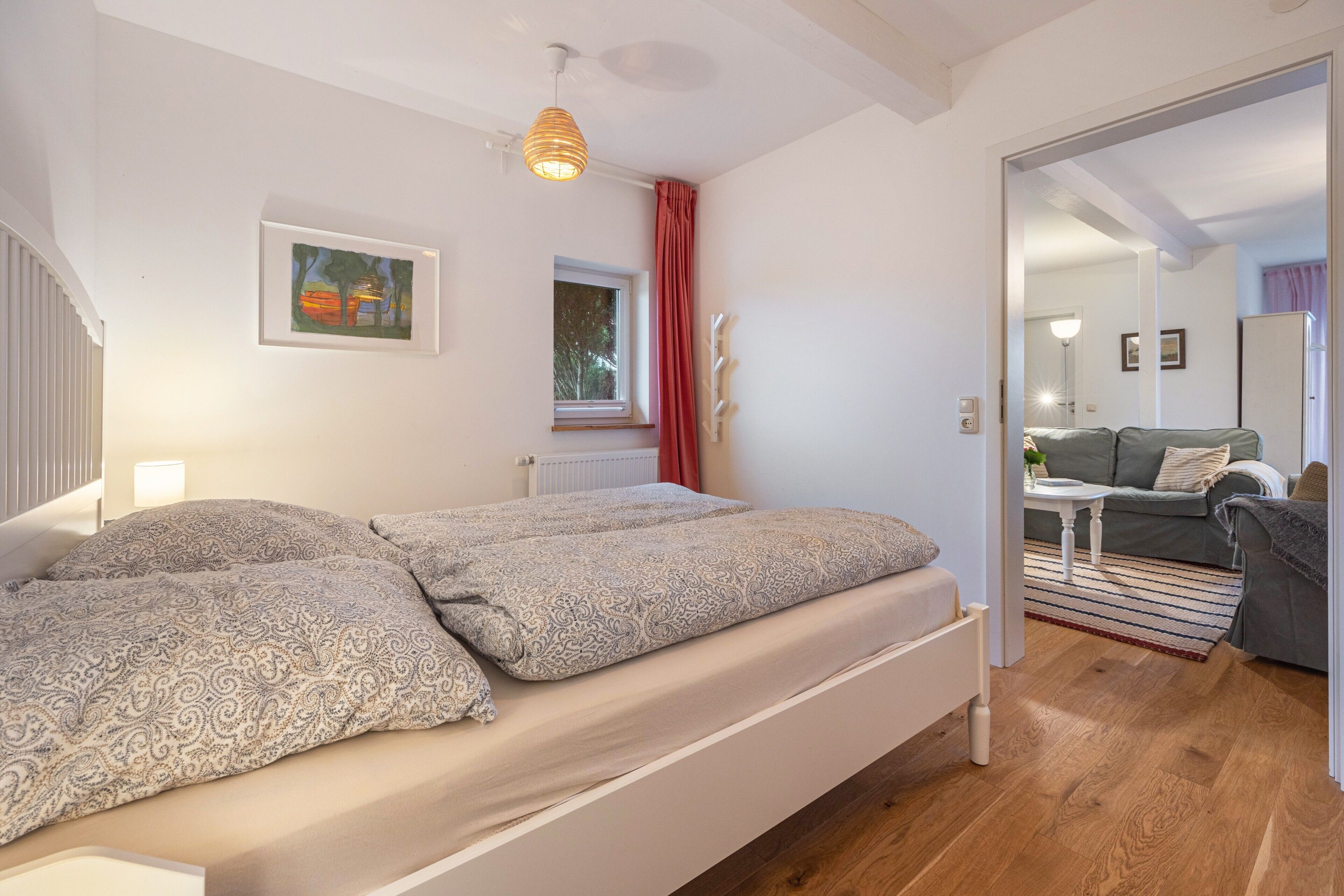 A bedroom with a bed, a window, a radiator, a door leading to another room, and a painting on the wall.