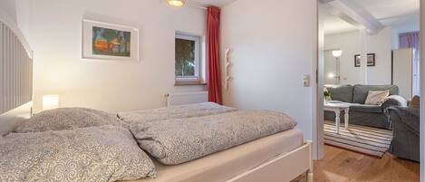 A bedroom with a bed, a window, a radiator, a door leading to another room, and a painting on the wall.