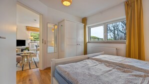 Une chambre à coucher comprenant un lit, une armoire, une fenêtre avec des rideaux, un radiateur et un téléviseur situé dans la pièce adjacente.