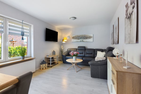 Ein modernes Wohnzimmer mit Sofa, Couchtisch, Fernseher und einem Fenster mit Blick auf Grünflächen.